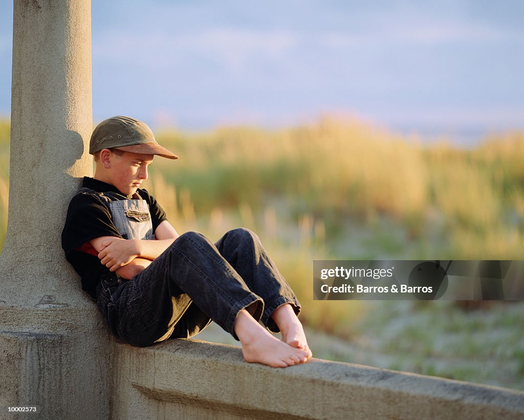 PENSIVE, BAREFOOT BOY SITTING IN OVERALLS