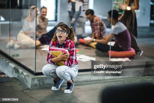 shocked nerdy student crouching with books at campus. - fish out of water stock pictures, royalty-free photos & images