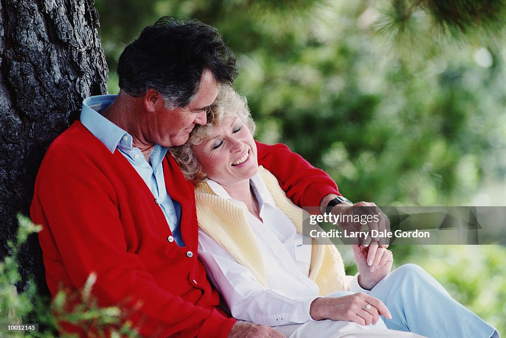 MIDDLE-AGED COUPLE RELAXING AGAINST TREE