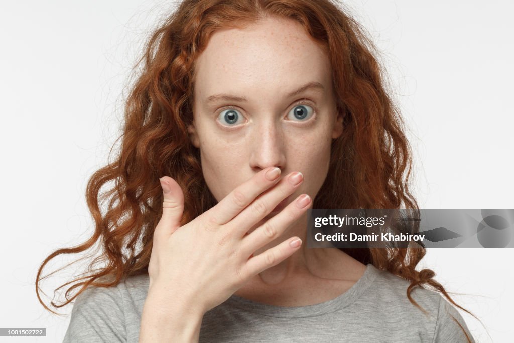 Astonished bug-eyed curly redhead girl covering her mouth with one hand, looking shocked. Surprised, embarrassed and confused female showing omg emotion