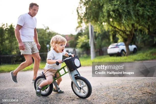 a small toddler boy with his grandfather riding a balance bike outdoors. - boyshorts fotografías e imágenes de stock
