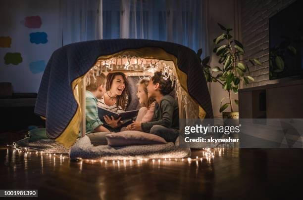 young cheerful nanny reading stories to small kids in a tent at home. - nanny stock pictures, royalty-free photos & images