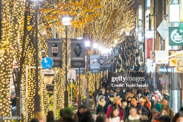 a crowd of people walks down the sidewalk beside the omotesando street among the illuminated row of zelkova trees and many fashion boutiques for winter holydays season in the night at jingumae, shibuyatokyo japan on december 06 2017. - harajuku tokyo harajuku street fashion stock pictures, royalty-free photos & images