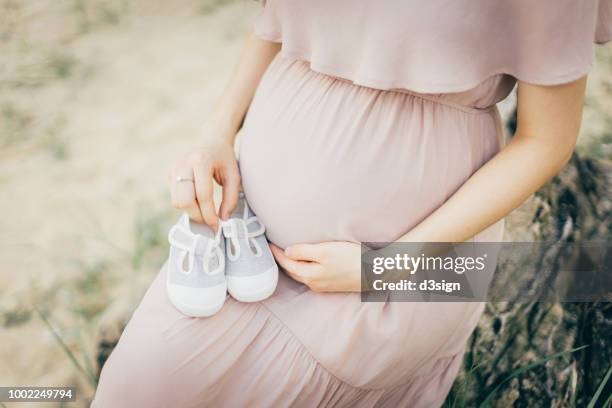 close up of pregnant woman wearing pink dress holding bump and baby shoes in nature park - pregnant belly stock pictures, royalty-free photos & images