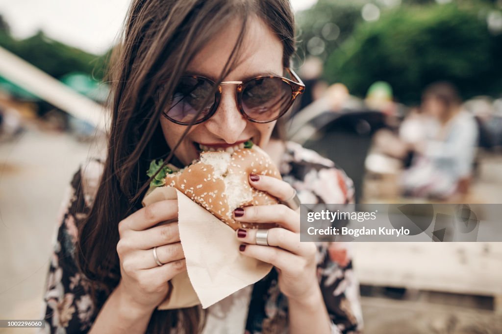 Stylish hipster woman eating juicy burger. boho girl biting yummy cheeseburger, smiling at street food festival. summertime. summer vacation travel picnic. space for text