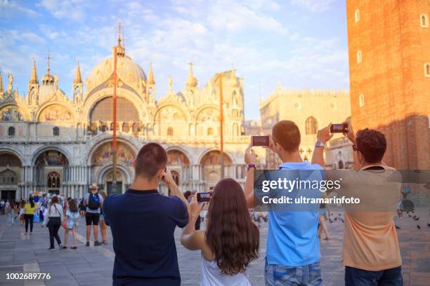 tourists in venezia. young women and young men, have fun. group of friends taking a  photo with smartphone, st mark's cathedral in piazza san marco, venice. casual lifestyles urban scene italy.  visiting venice, italy. - saint mark stock pictures, royalty-free photos & images