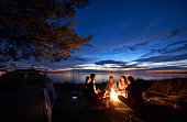 Night summer camping on shore. Group of young tourists around campfire near tent under evening sky