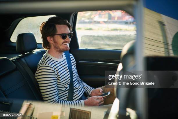 smiling man with mobile phone sitting in backseat of car - car passenger stock pictures, royalty-free photos & images