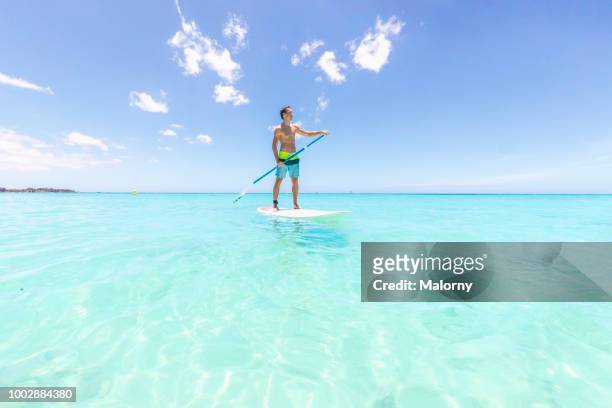 young man standing on a sup board or paddleboard, floating on the clear blue water, looking away. trou-aux-biches, pamplemousses district, mauritius. - mauritius stock-fotos und bilder