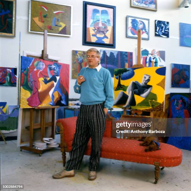 Portrait of English painter David Hockney, dressed in a light blue sweatshirt and tan trousers, as he stands in front of a chaise longue in his home,...