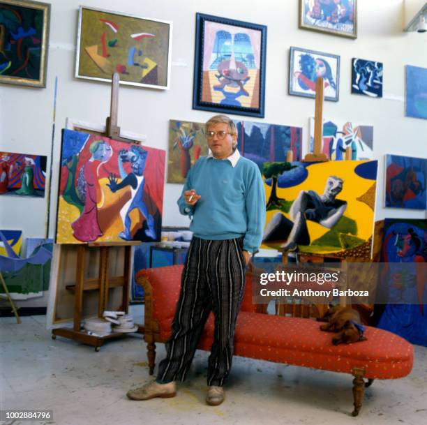 Portrait of English painter David Hockney, dressed in a light blue sweatshirt and tan trousers, as he stands in front of a chaise longue in his home,...