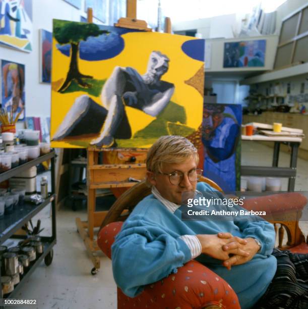 Portrait of English painter David Hockney, dressed in a light blue sweatshirt and tan trousers, as he sits on a chaise longue in his home, Los...