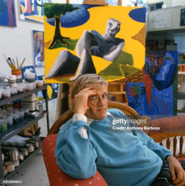 Portrait of English painter David Hockney, dressed in a light blue sweatshirt and tan trousers, as he sits on a chaise longue in his home, Los...