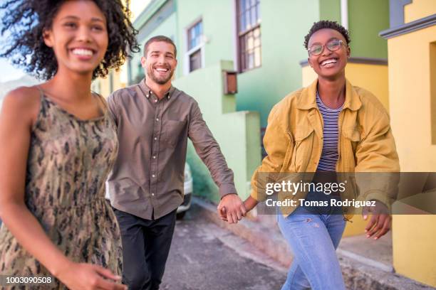 three happy friends hand in hand on city street - man running city stock-fotos und bilder