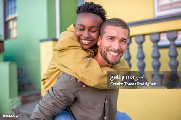 portrait of happy young man giving girlfriend a piggyback ride - multiracial couple stock-fotos und bilder