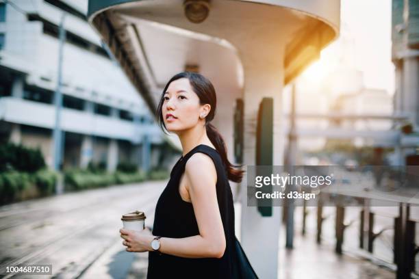 beautiful young lady holding coffee cup waiting for tram at station in city - waiting bus stock pictures, royalty-free photos & images