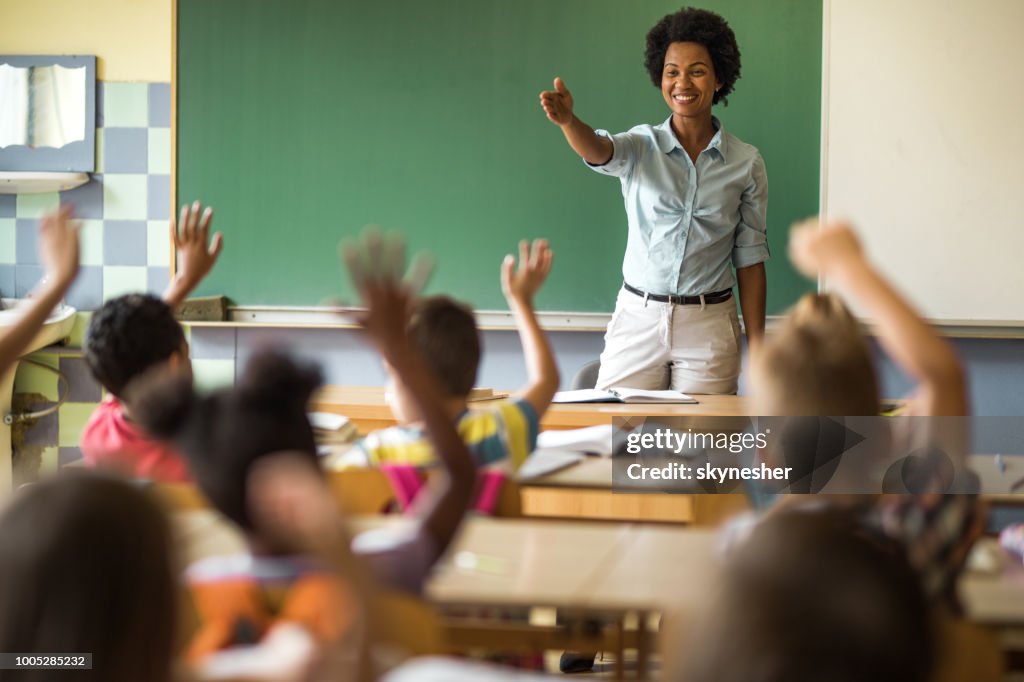 Happy African American elementary teacher aiming at school kid to answer her question.