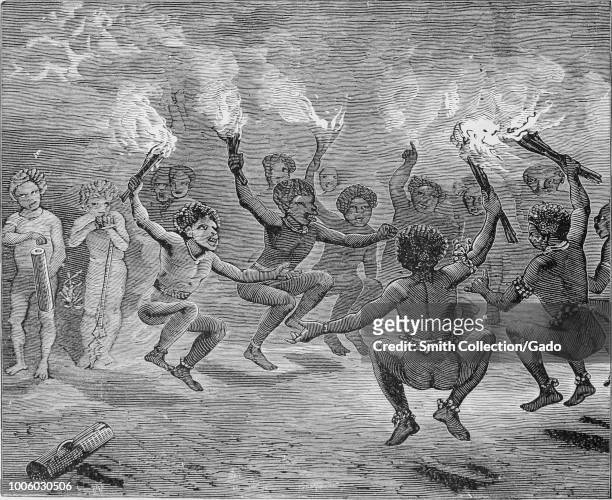 Black and white print depicting a group of New Guinean men holding lit torches and dancing in a circle while other figures look on from the...