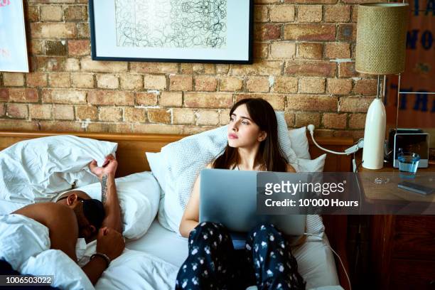 pensive young woman using laptop in bed with partner asleep - african american man day dreaming stock pictures, royalty-free photos & images