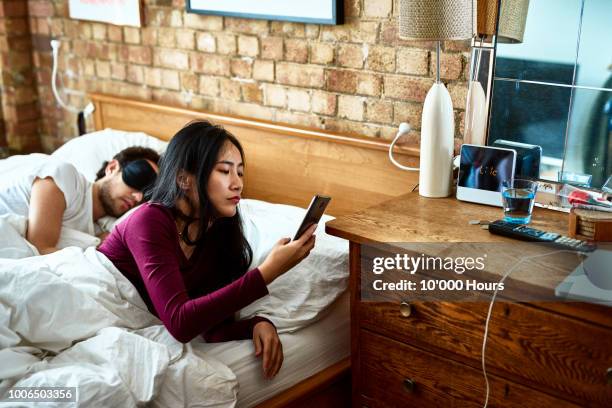 chinese woman checking smartphone as man sleeps beside in bed - kommunikationsproblem stock-fotos und bilder