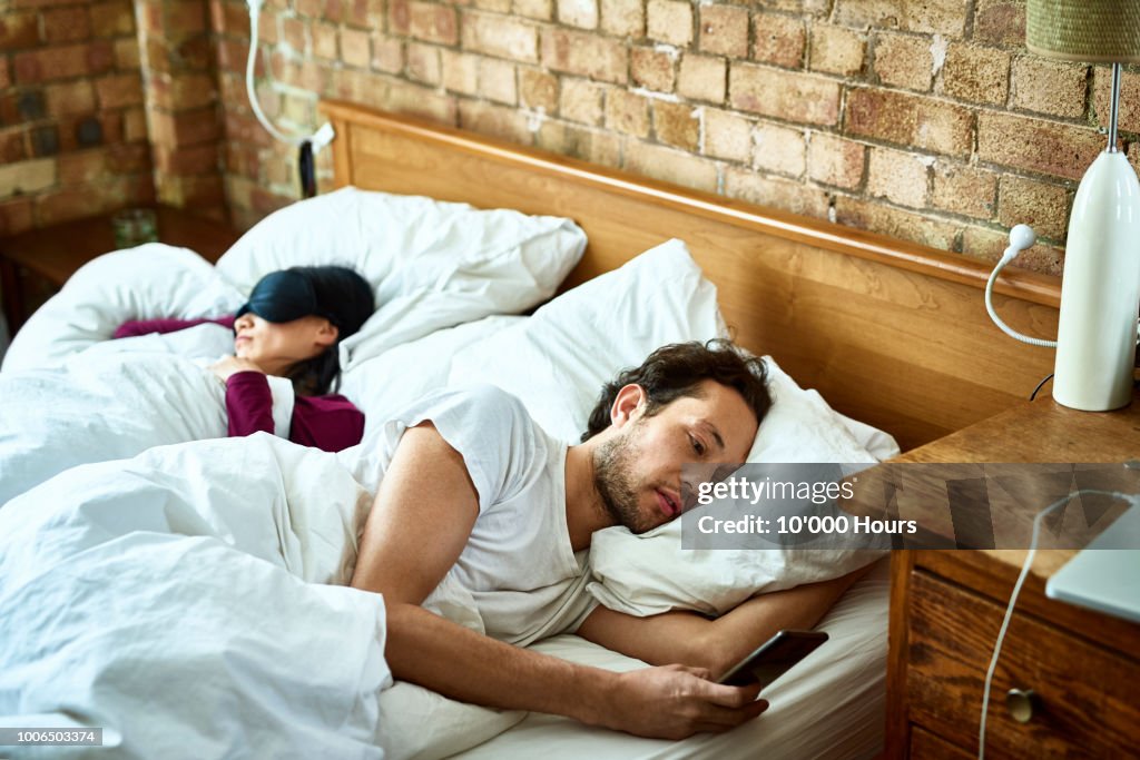 Woman fast asleep next to partner who is checking his smartphone