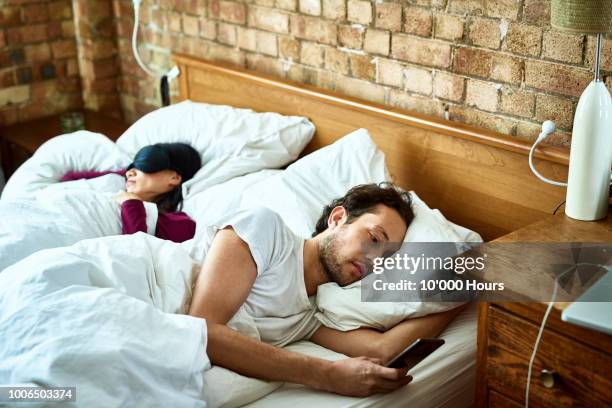 woman fast asleep next to partner who is checking his smartphone - affari stock pictures, royalty-free photos & images