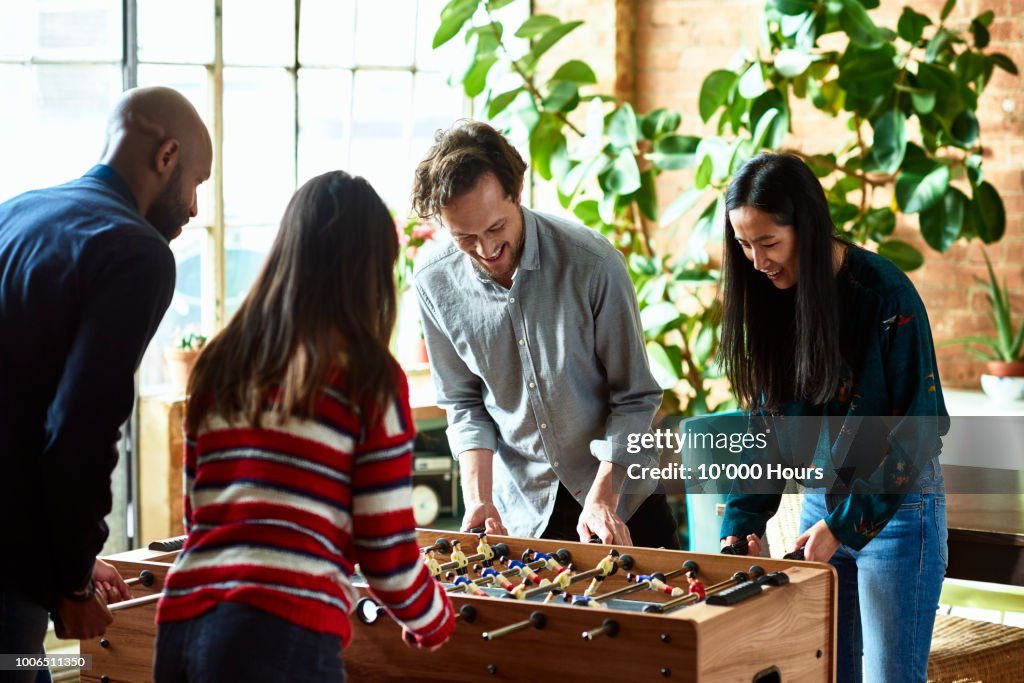Four friends playing foosball and laughing in modern apartment