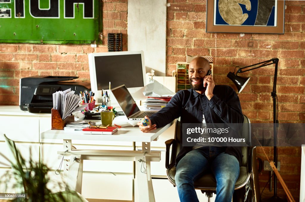 Cheerful casual businessman sitting at desk on phone and smiling