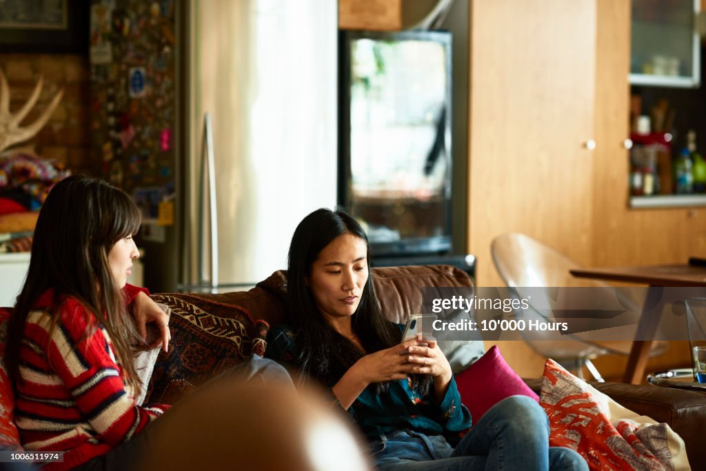 Chinese woman sitting on sofa with friend using cell phone