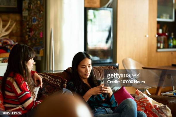 chinese woman sitting on sofa with friend using cell phone - friends arguing stock pictures, royalty-free photos & images