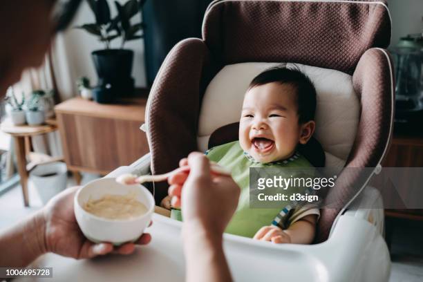 father feeding smiling happy baby solid food with spoon on high chair at home - asian baby eating fotografías e imágenes de stock