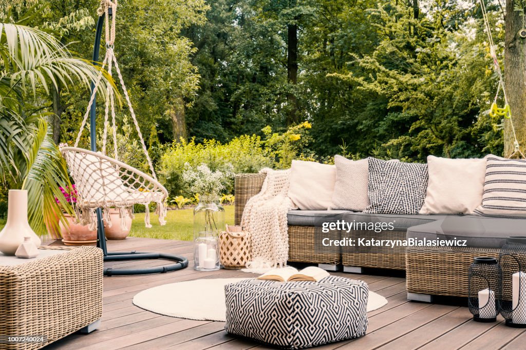 Book on a black and white pouf in the middle of a bright terrace with a rattan corner sofa, hanging chair and round rug. Real photo