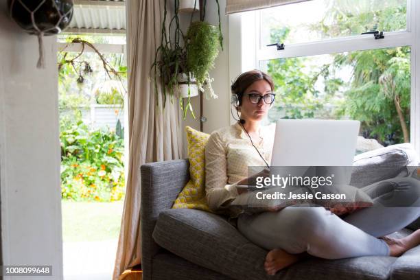 woman working from home sitting on a sofa working on a laptop wearing a headset - new zealand business stock pictures, royalty-free photos & images
