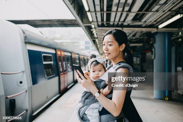 Young mother with baby girl using smartphone while waiting for subway in subway station platform