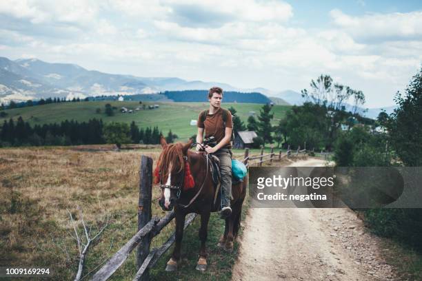 smiling young man in the brown t-shirt with green backpack and binoculars sits on the back of the brown horse in the mountains - riding hat stock-fotos und bilder