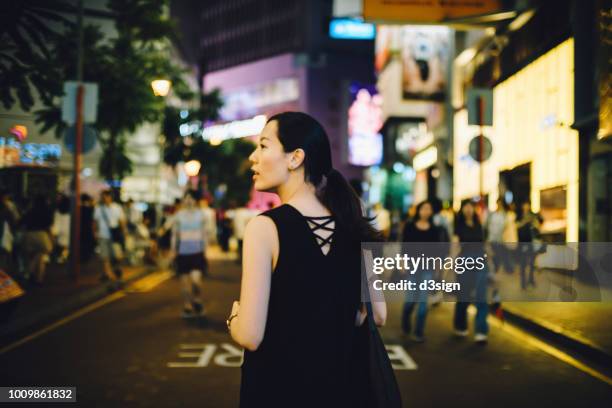 rear view of young woman strolling down city street looking sideways at night - causeway bay stock pictures, royalty-free photos & images