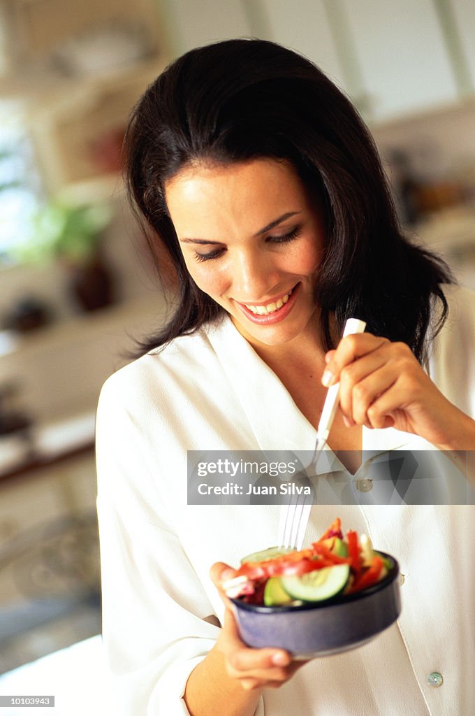 WOMAN EATING SALAD FROM BOWL IN KITCHEN
