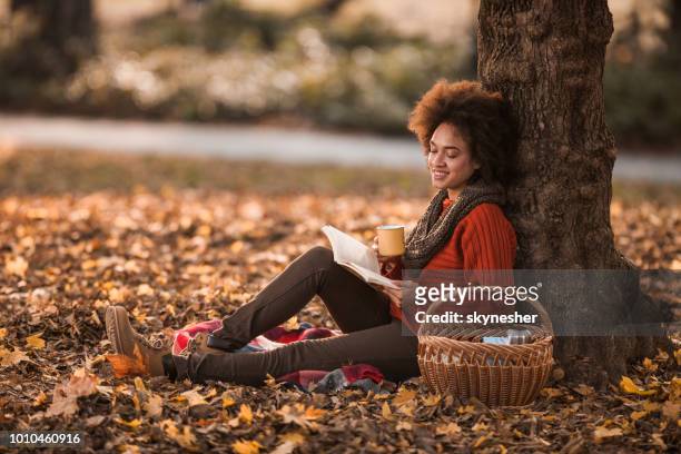 happy black woman reading a book while enjoying on picnic in autumn day. - autumn coffee stock pictures, royalty-free photos & images