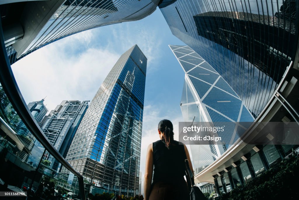 Rear view of professional young businesswoman standing against contemporary financial skyscrapers in downtown financial district and looking up into sky with positive emotion