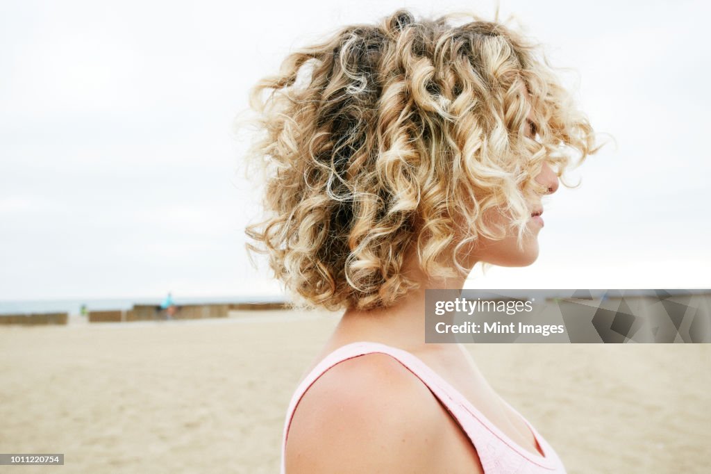 Profile portrait of young woman with blond curly hair standing on sandy beach.