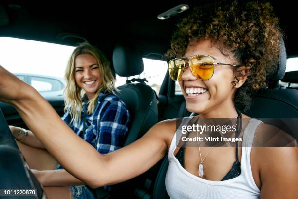 portrait of two women with long blond and brown curly hair sitting in car, wearing sunglasses, smiling. - automobile and fun stockfoto's en -beelden