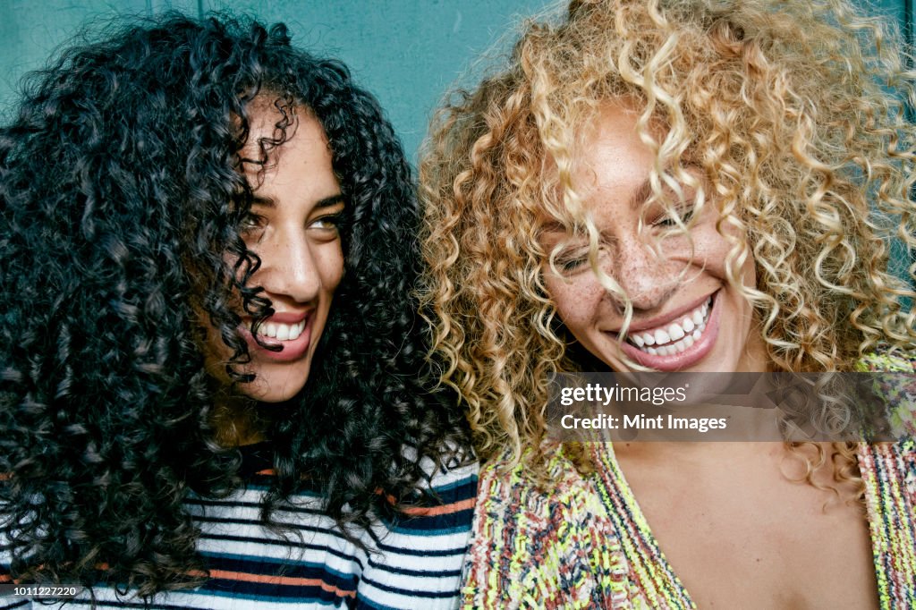 Portrait of two young women with long curly black and blond hair, smiling and laughing.