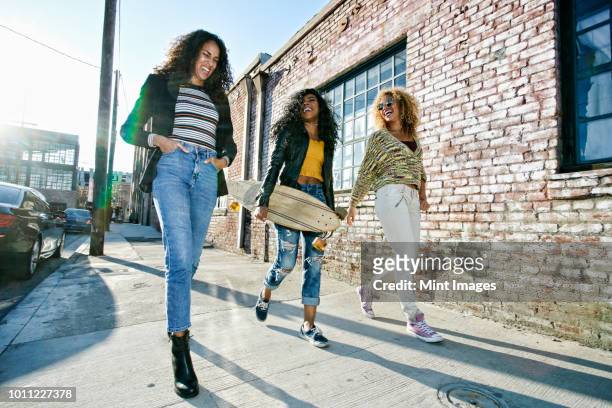 three young women with long curly hair walking along pavement, one carrying skateboard. - city of los angeles stockfoto's en -beelden