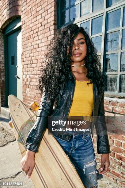 young smiling woman with long curly black hair standing on pavement, holding skateboard, looking at camera. - bauchfreies oberteil stock-fotos und bilder