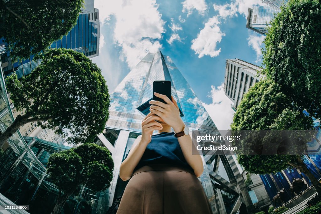 Confident young businesswoman using mobile phone in front of highrise financial towers in Central Business District on a fresh bright morning