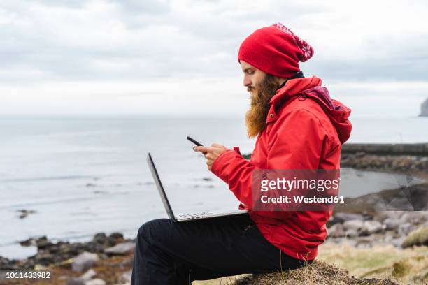 iceland, north of iceland, man with laptop sitting near the sea using cell phone - le cap - fotografias e filmes do acervo