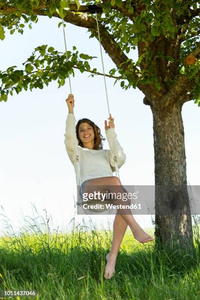 smiling young woman sitting on swing - frau wiese bewegt stock-fotos und bilder