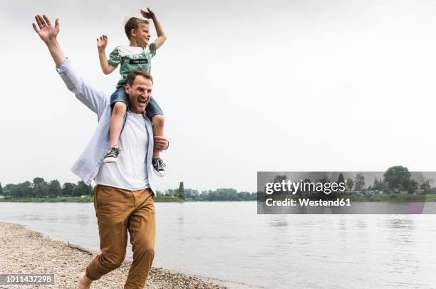 happy father carrying son on shoulders at the riverside - mann mit kind auf den schultern stock-fotos und bilder