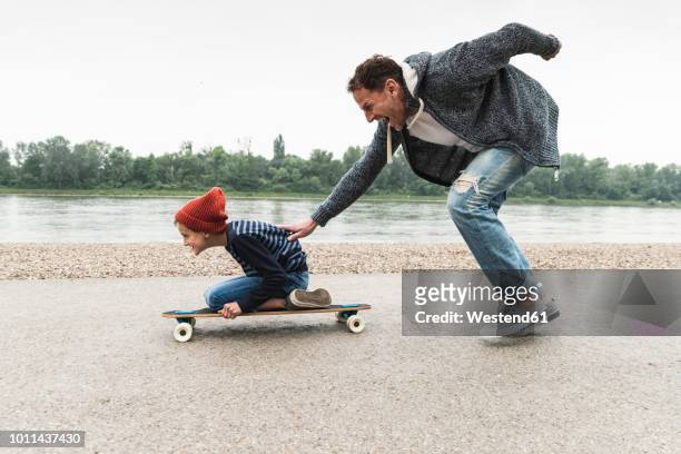 happy father pushing son on skateboard at the riverside - in movimento foto e immagini stock