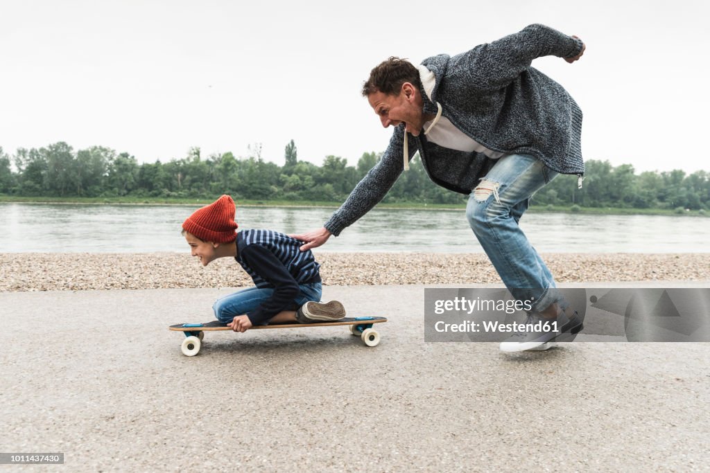 Happy father pushing son on skateboard at the riverside